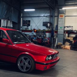 RAJAUTO CENTER ANDRZEJ MAGACZEWSKI - Czerwone BMW E30 z charakterystycznymi felgami w warsztacie samochodowym z widocznymi mechanikami w tle. Auto po renowacji/tuningu.