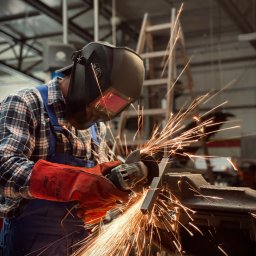 RAJAUTO CENTER ANDRZEJ MAGACZEWSKI - Spawanie metalu w warsztacie: spawacz w masce ochronnej, iskry rozbryzgują się podczas pracy. W tle drabina i konstrukcja dachu.