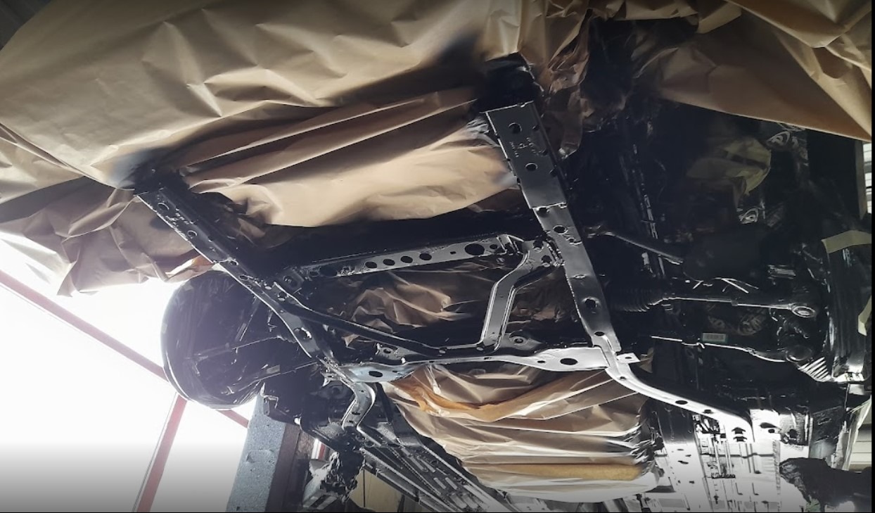 Podwozie samochodu po konserwacji antykorozyjnej, zabezpieczone papierem maskującym, widoczne elementy zawieszenia i układu kierowniczego pomalowane na czarno.