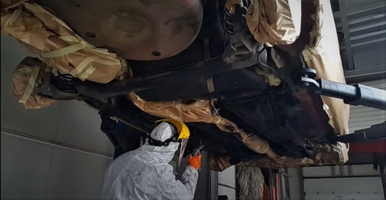 Pracownik w kombinezonie ochronnym, z maską i rękawicami, piaskuje podwozie samochodu na podnośniku. Elementy auta zabezpieczone papierem i taśmą.