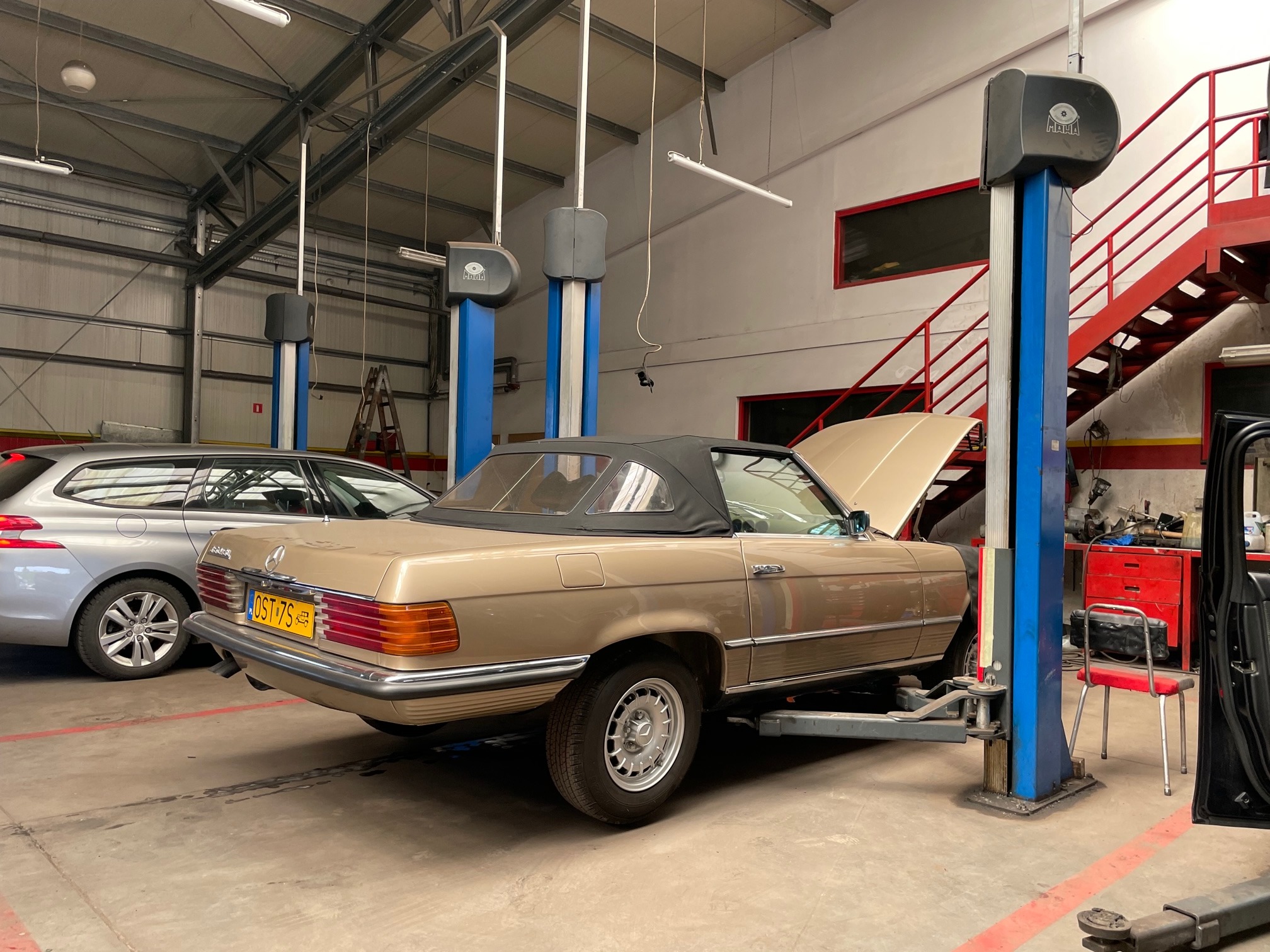 Beżowy kabriolet Mercedes-Benz na podnośniku w warsztacie. Otwarta maska, widoczny numer rejestracyjny, obok szary samochód osobowy, w tle schody i narzędzia.