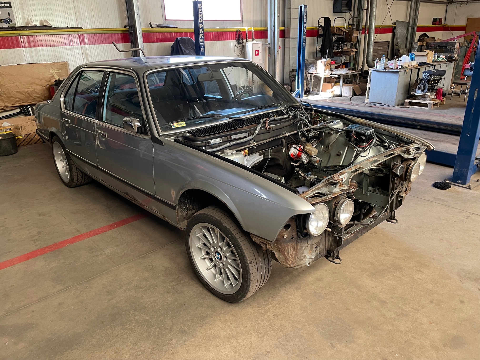 Szary, klasyczny samochód BMW bez maski w warsztacie, widoczne podnośniki. Silnik w trakcie naprawy, eksponowane przewody i elementy mechaniczne.