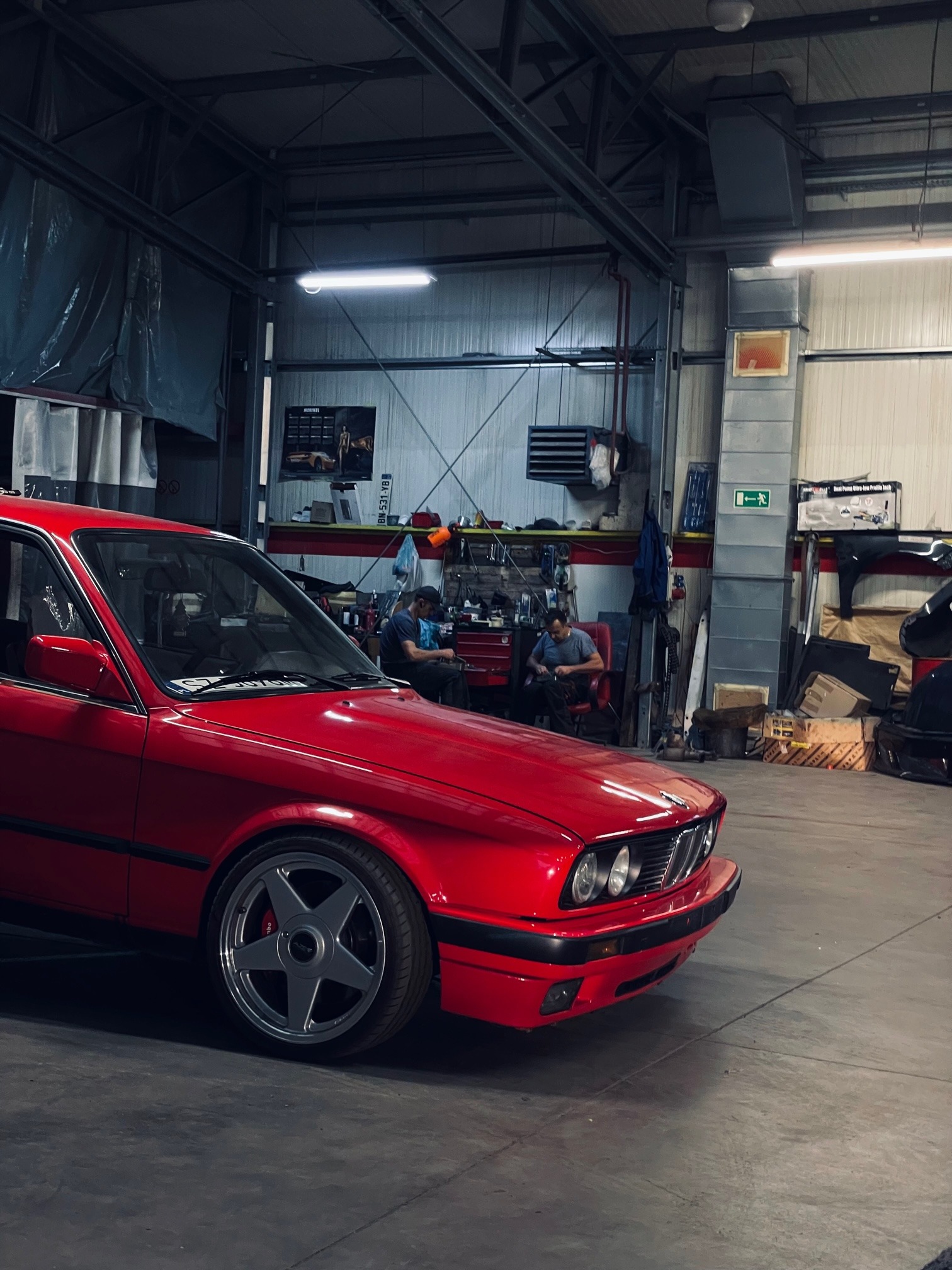 Czerwone BMW E30 z charakterystycznymi felgami w warsztacie samochodowym z widocznymi mechanikami w tle. Auto po renowacji/tuningu.