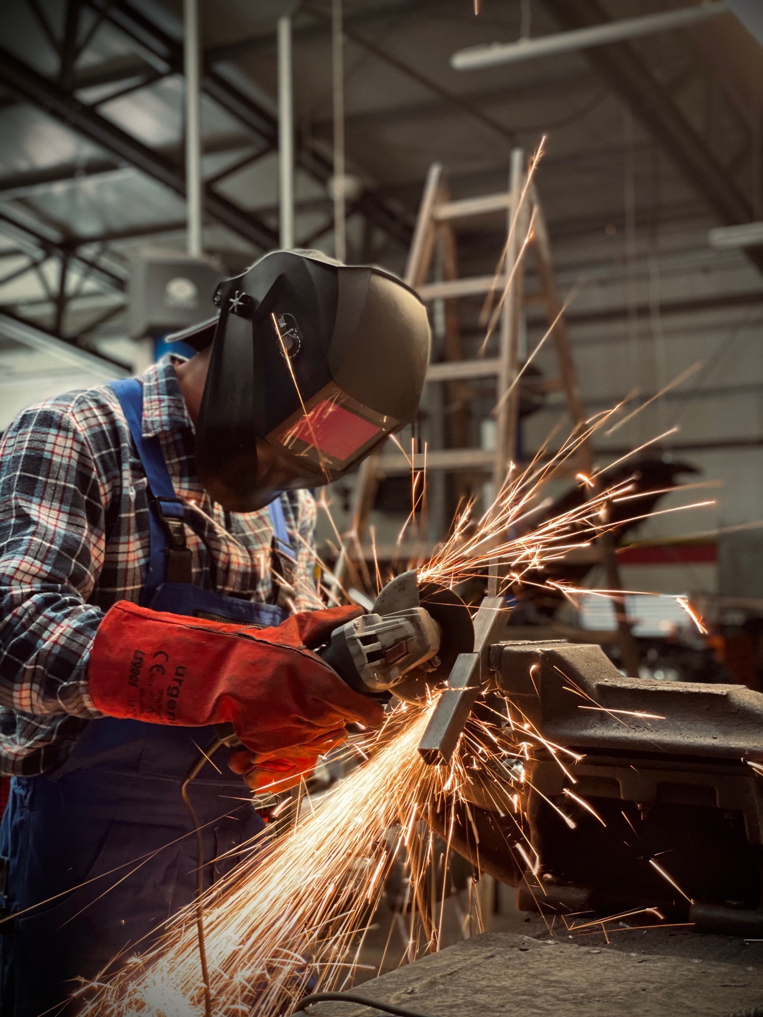 Spawanie metalu w warsztacie: spawacz w masce ochronnej, iskry rozbryzgują się podczas pracy. W tle drabina i konstrukcja dachu.