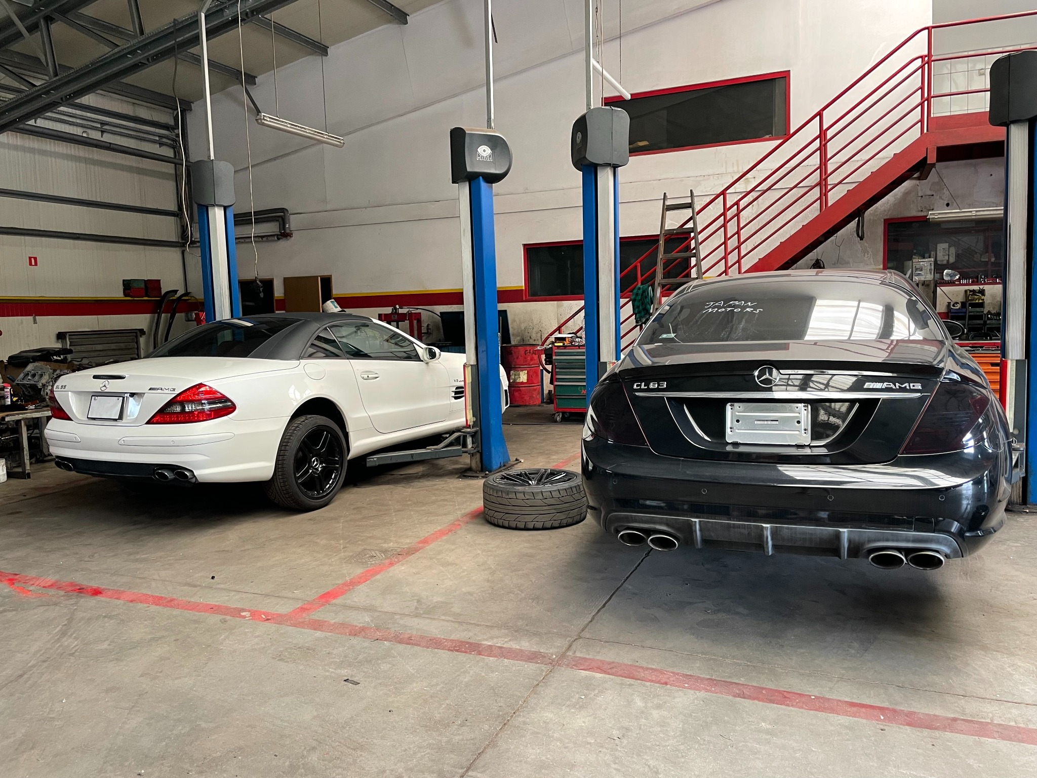 Dwa auta Mercedes-Benz (biały SL i czarny CLS) w warsztacie samochodowym, na podnośnikach. Widoczne narzędzia i wyposażenie warsztatu, w tym opony.