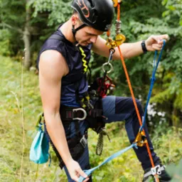Arborysta w uprzęży i kasku, przygotowuje się do pracy na wysokości w otoczeniu zieleni. Widoczne liny, karabinki i specjalistyczny sprzęt.