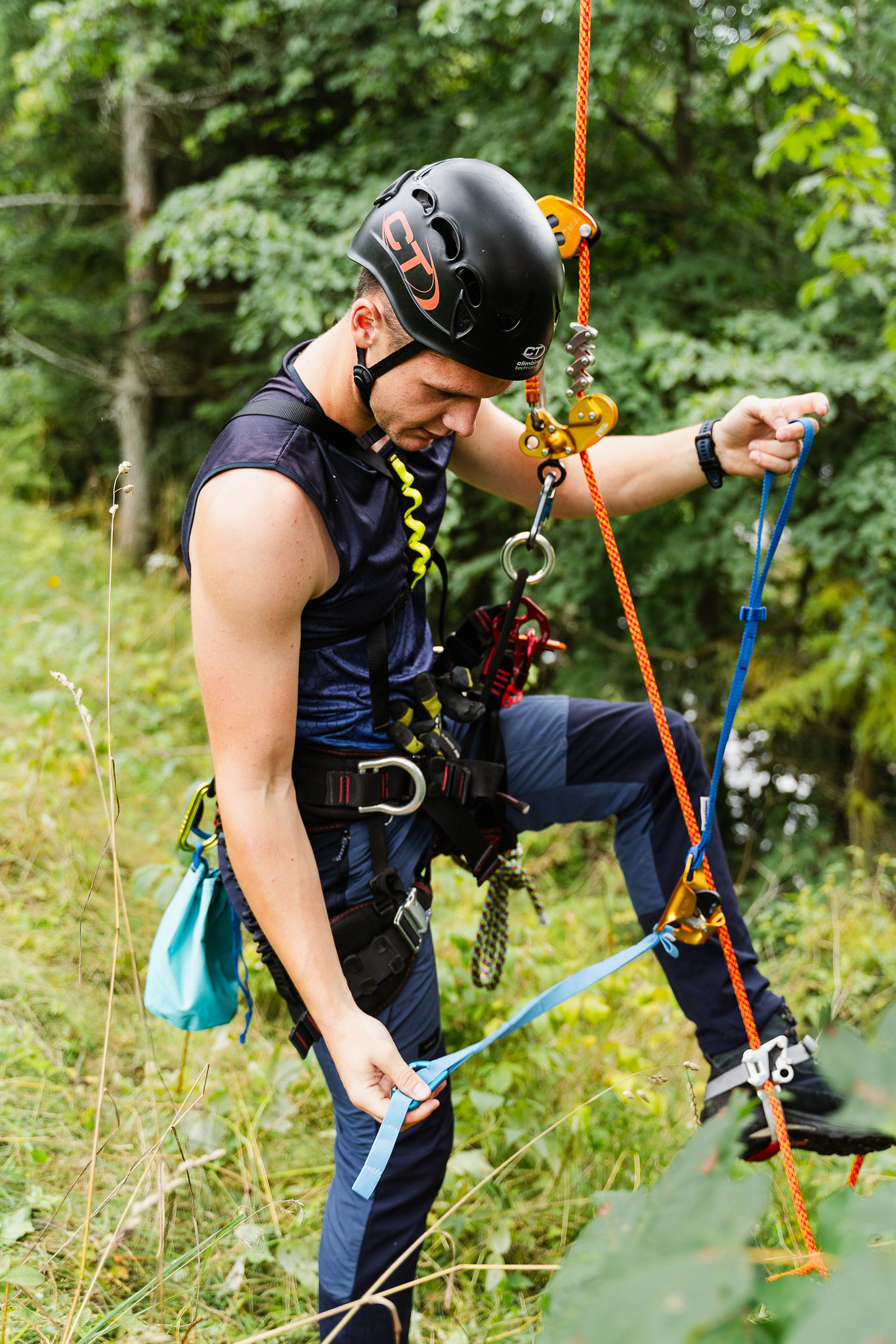 Arborysta w uprzęży i kasku, przygotowuje się do pracy na wysokości w otoczeniu zieleni. Widoczne liny, karabinki i specjalistyczny sprzęt.
