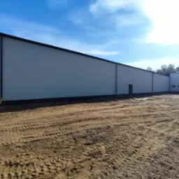 Nowoczesny budynek użyteczności publicznej z płaskim dachem i elewacją w kolorze jasnej szarości, widoczny z poziomu gruntu na tle błękitnego nieba.