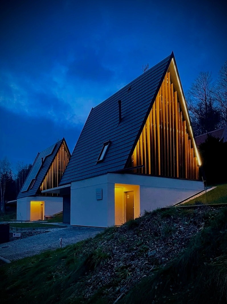 Dwa nowoczesne domki letniskowe oświetlone od wewnątrz, z charakterystycznymi trójkątnymi dachami i drewnianą elewacją, sfotografowane o zmierzchu w Radkowie.