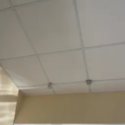 Kasetonowy sufit podwieszany z czujnikiem i puszką instalacyjną. Widok z dołu na białe płyty i szarą konstrukcję nośną.