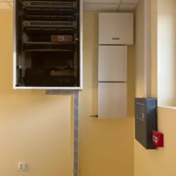 Szafa rack, panele Kade i centrala alarmowa na żółtej ścianie. Widoczne okablowanie i gniazdko elektryczne. Instalacja systemów bezpieczeństwa w biurze.