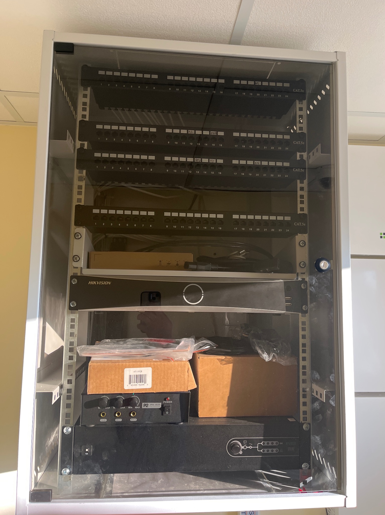 Szafa rack z patch panelem CAT5e, rejestratorem Hikvision, wzmacniaczem i akcesoriami. Widoczne kable i złącza. Zabezpieczona zamkiem.