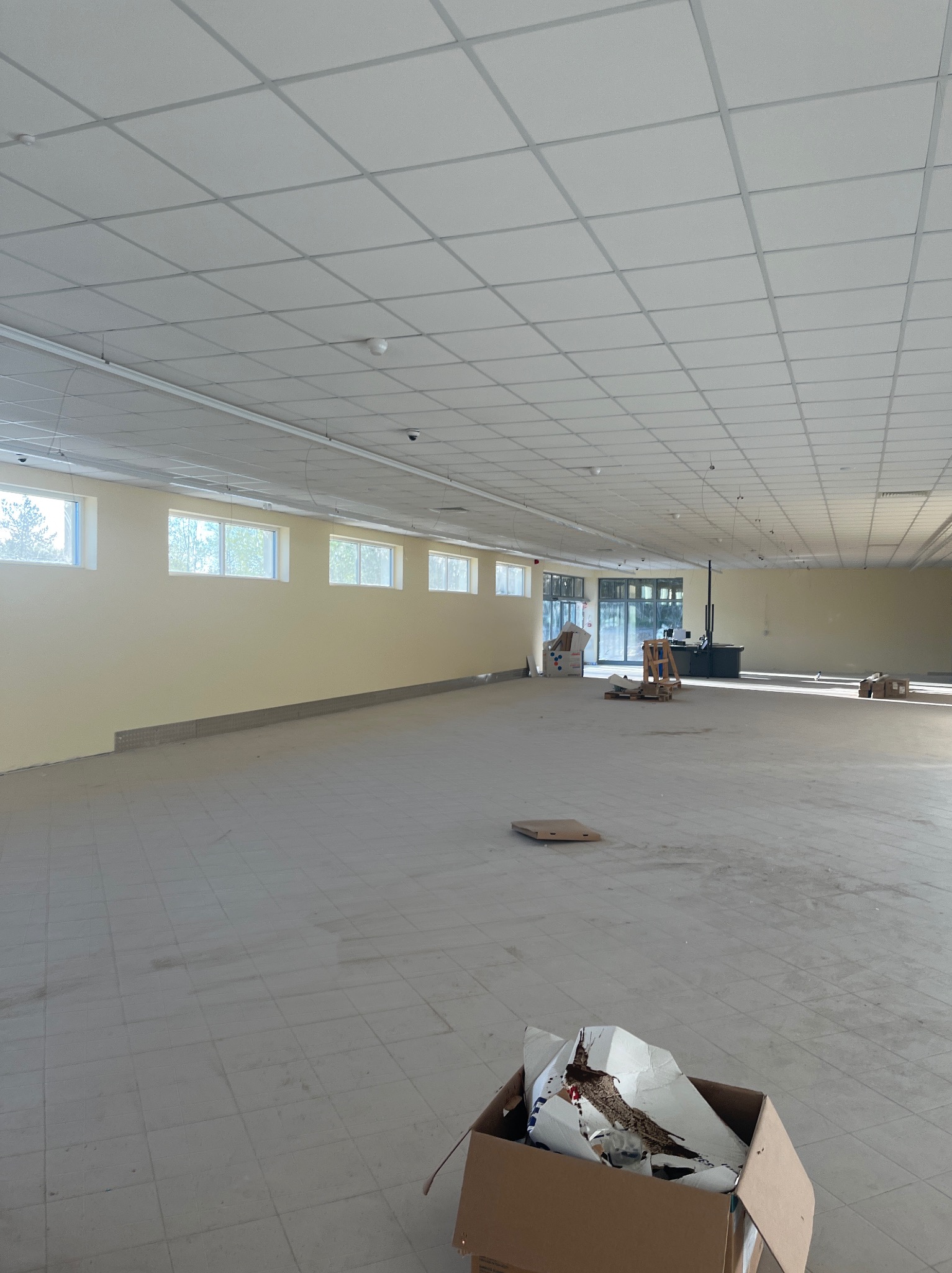 Przestronne wnętrze hali z oknami, sufit podwieszany z oświetleniem. Widoczne elementy wyposażenia i kartony. Hala w trakcie prac wykończeniowych.
