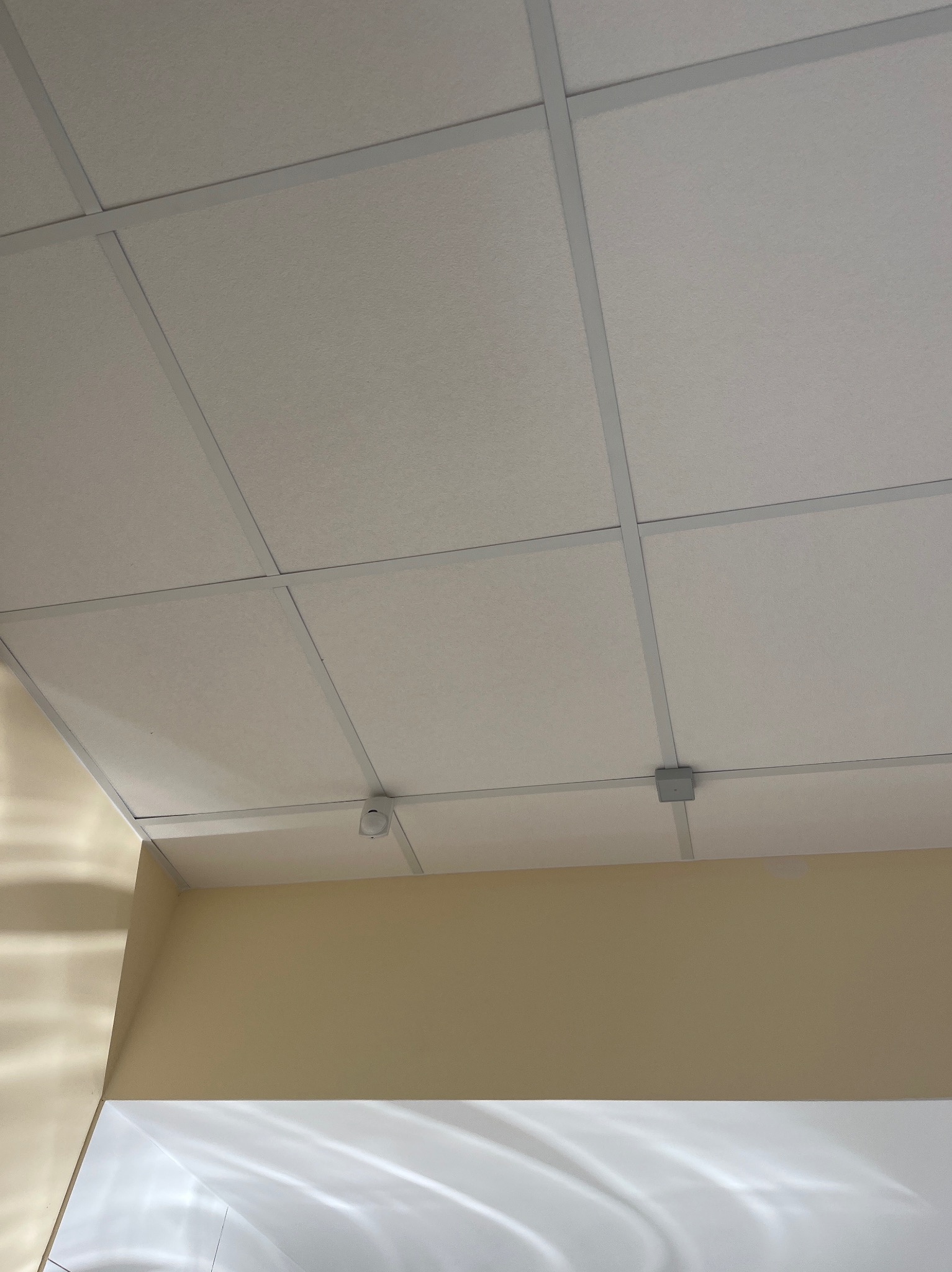 Kasetonowy sufit podwieszany z czujnikiem i puszką instalacyjną. Widok z dołu na białe płyty i szarą konstrukcję nośną.