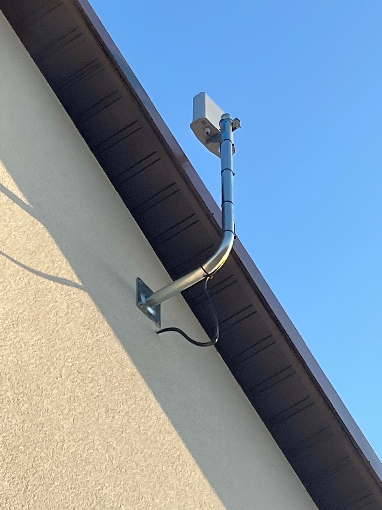 Zewnętrzny montaż kamery monitoringu na elewacji budynku, widok z dołu na błękitne niebo. Metalowa rura mocująca z kablem. Kremowa elewacja i brązowe podbitki dachowe.