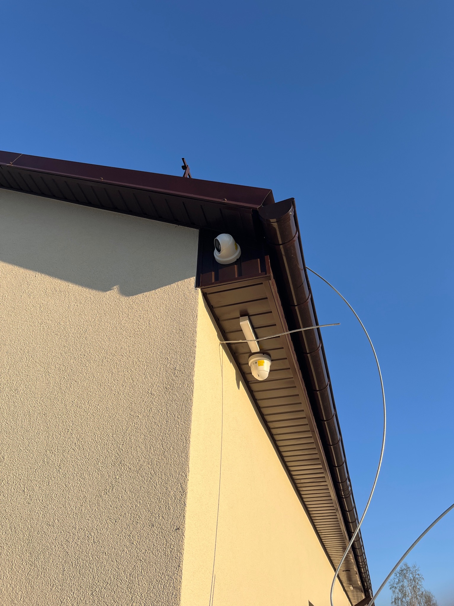 Dwie kamery monitoringu zamontowane na elewacji budynku z brązowym dachem, widoczne kable i fragment drzewa na tle błękitnego nieba. Ujęcie z dołu.