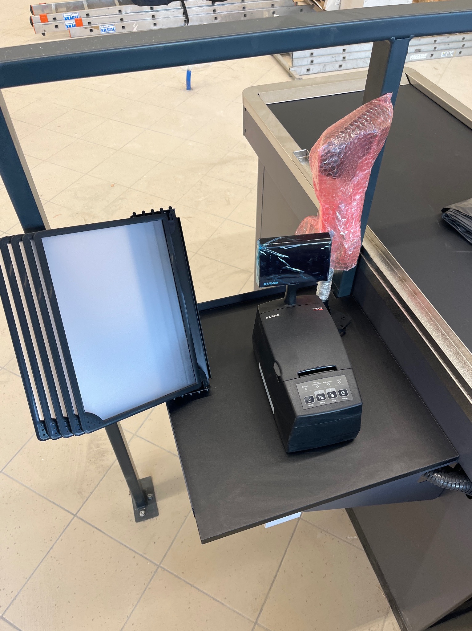 Stanowisko kasowe z czarnym terminalem płatniczym ELZAB i pustym, podświetlanym panelem informacyjnym, widoczne drabiny Krause w tle.