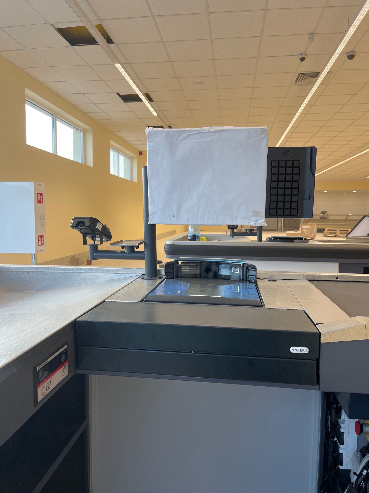 Kasa samoobsługowa w supermarkecie, z widocznymi elementami systemu monitoringu: kamerą pod sufitem i czytnikiem kodów kreskowych. Oświetlenie sufitowe.