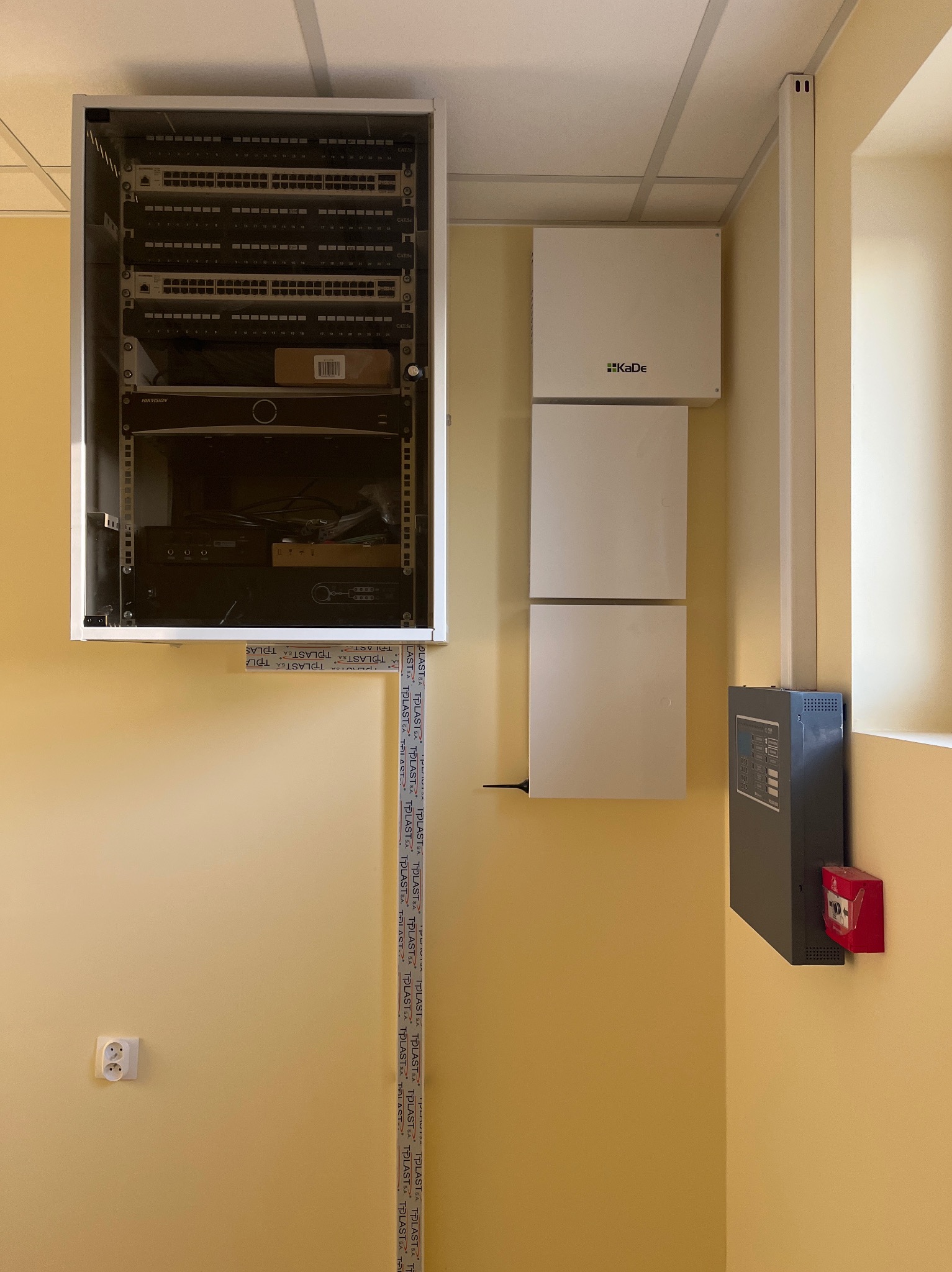 Szafa rack, panele Kade i centrala alarmowa na żółtej ścianie. Widoczne okablowanie i gniazdko elektryczne. Instalacja systemów bezpieczeństwa w biurze.