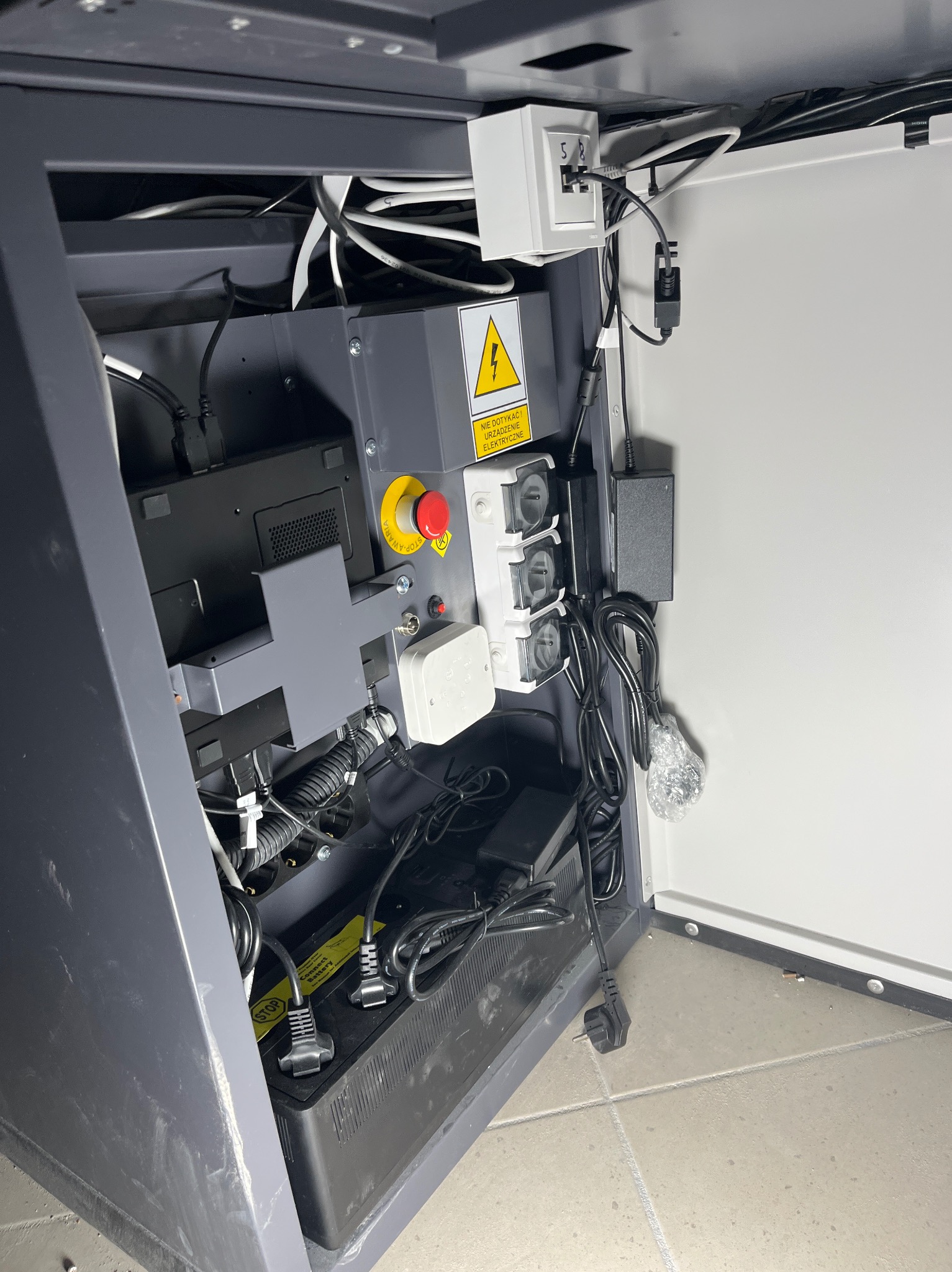 Wnętrze szafy sterowniczej: kable, zasilacze, gniazda, przycisk awaryjny i akumulator. Widoczne okablowanie i elementy elektryczne.