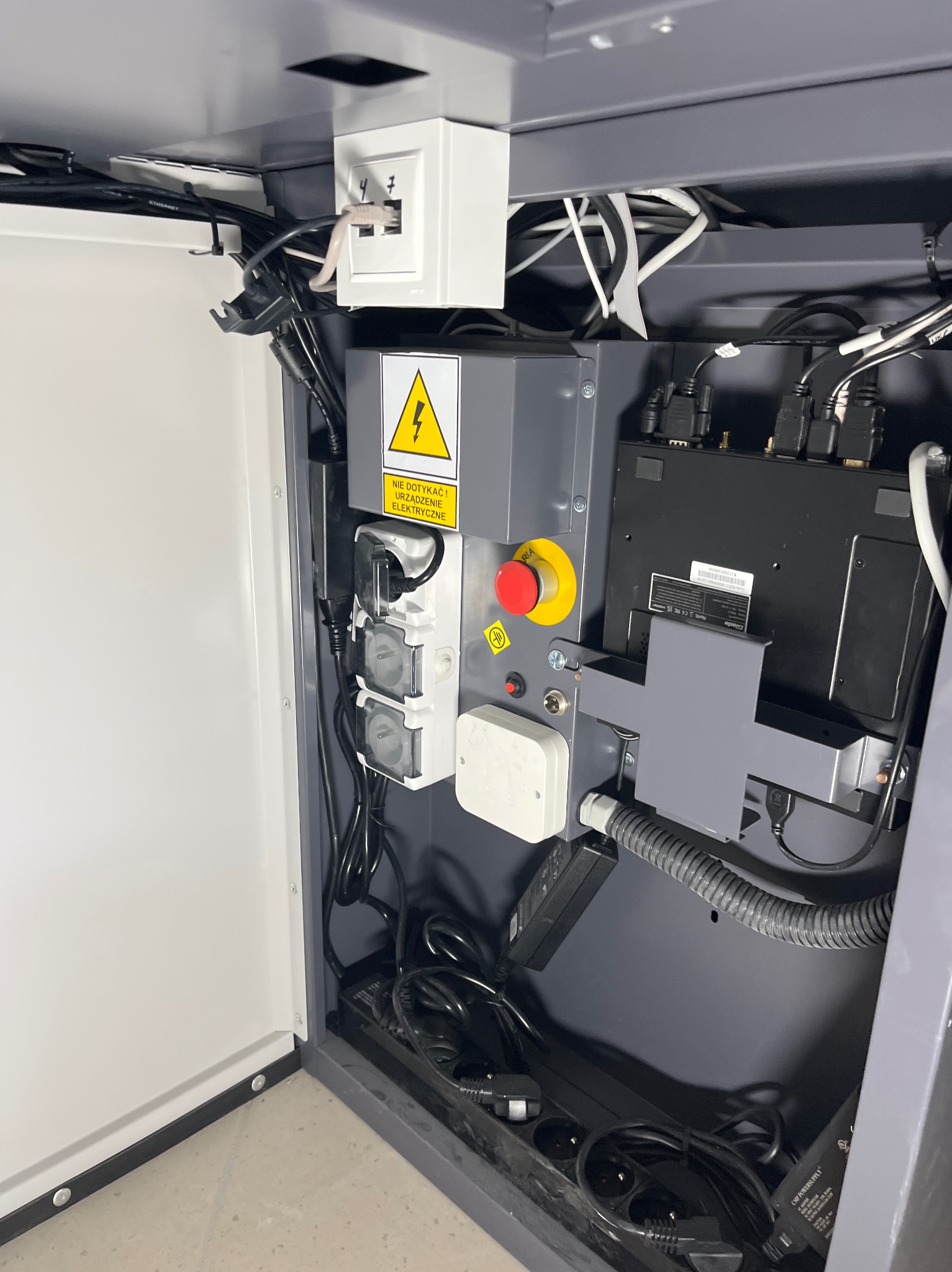 Szara szafka sterownicza z przyciskiem awaryjnym, gniazdami zasilającymi i licznymi kablami. Widoczna tabliczka ostrzegawcza o zagrożeniu elektrycznym.