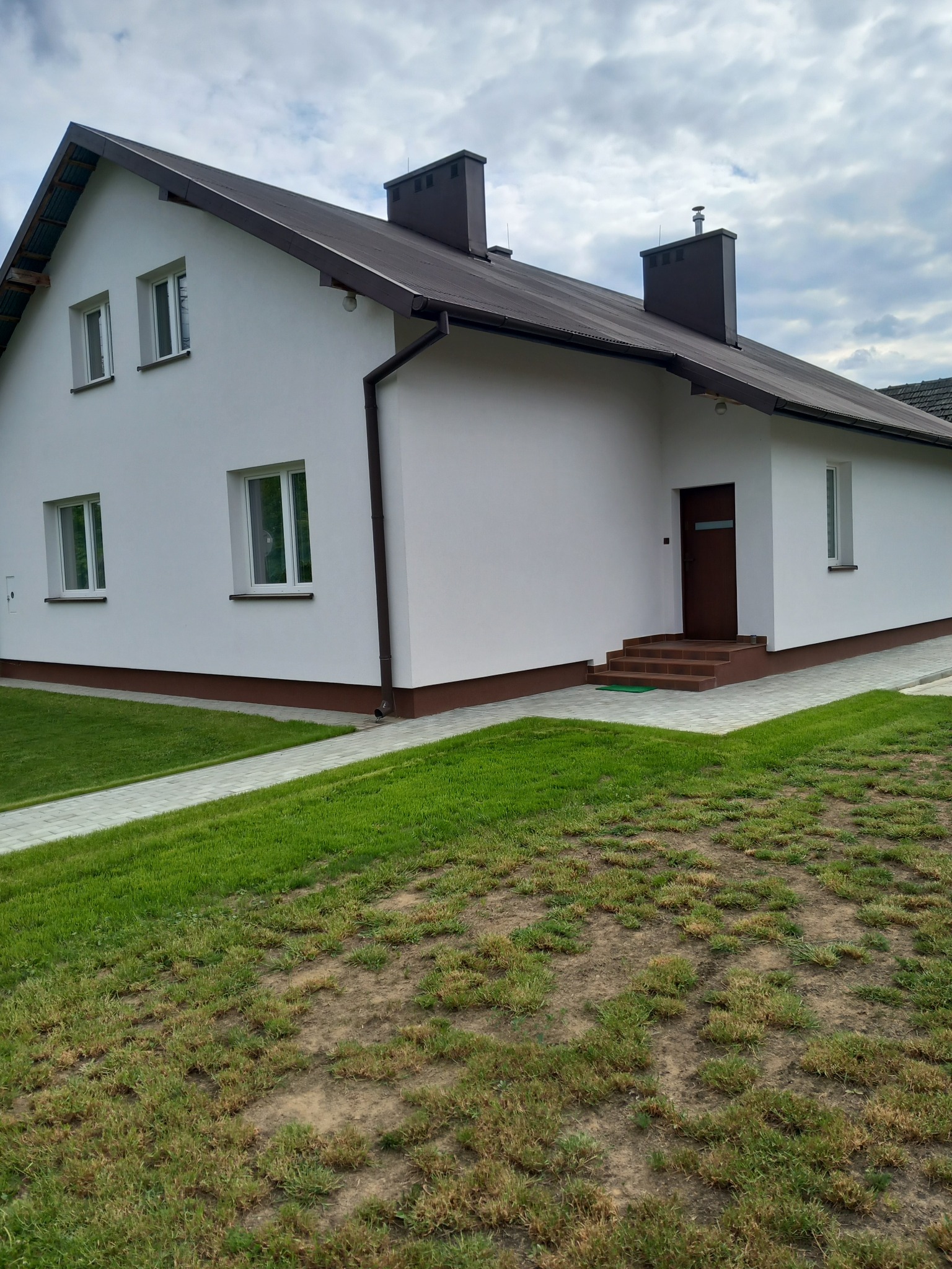 Biały dom jednorodzinny z brązowym dachem i elementami wykończeniowymi, widok z trawnika. Ścieżka z kostki prowadzi do wejścia.