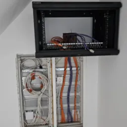 Rozdzielnica elektryczna z kablami w kolorach pomarańczowym, fioletowym i białym, zamontowana na ścianie, obok szafka rack z patch panelem.