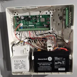 Otwarta obudowa centrali alarmowej z widoczną elektroniką, akumulatorem i transformatorem. Kable w różnych kolorach. Model Perfecta 16.