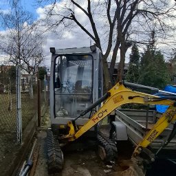 Vell Kom Jakub Kostrubiec - Żółto-szara mini koparka Wacker Neuson 1404 na gąsienicach, zaparkowana obok przyczepy na działce, z drzewami w tle i błękitnym niebem.