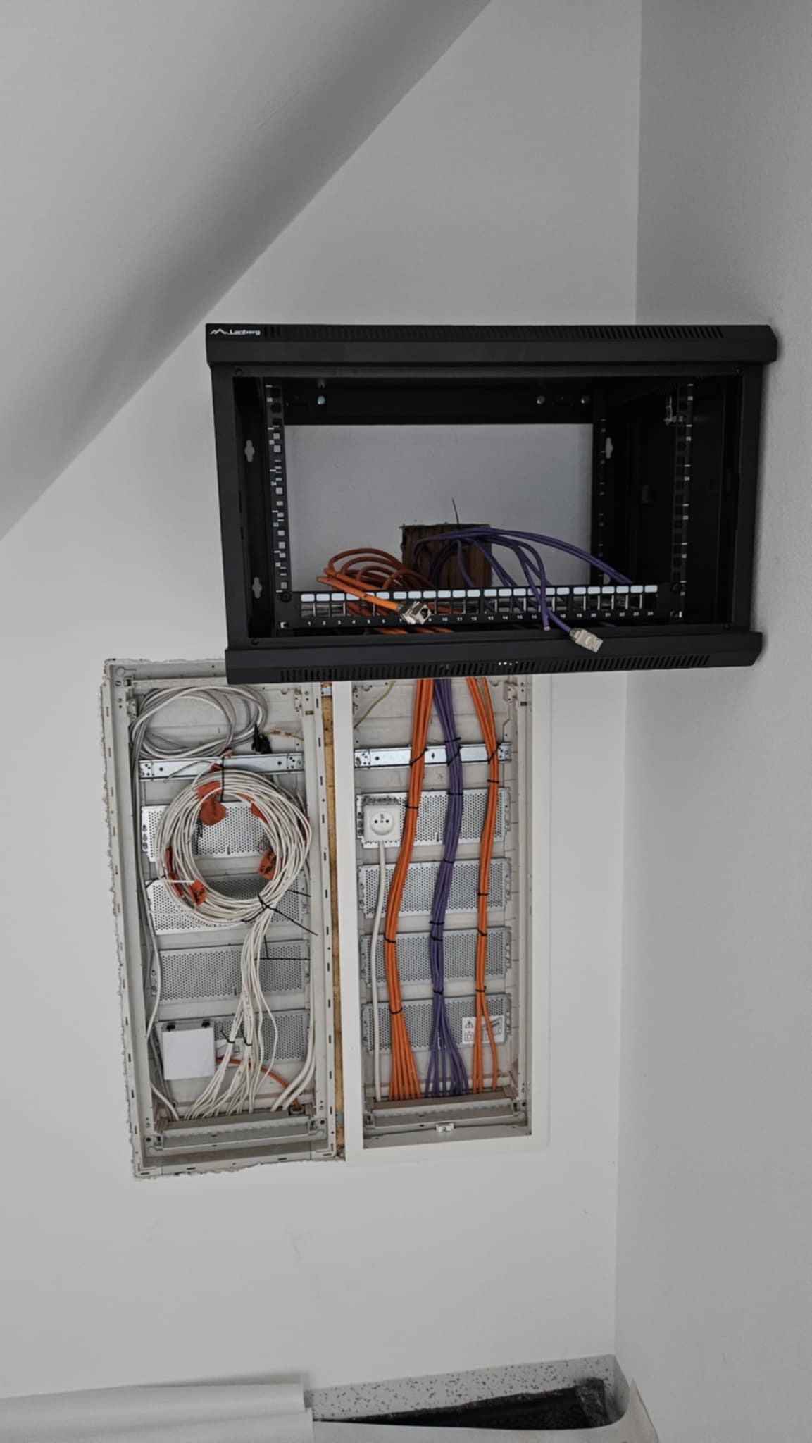 Rozdzielnica elektryczna z kablami w kolorach pomarańczowym, fioletowym i białym, zamontowana na ścianie, obok szafka rack z patch panelem.