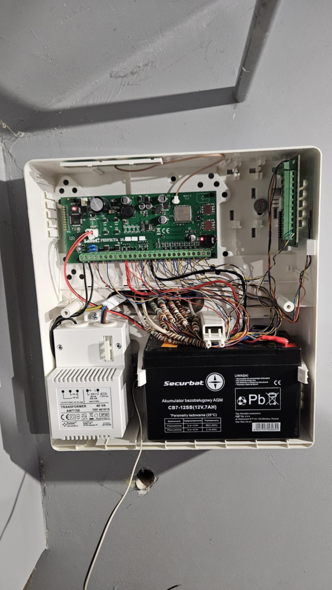 Otwarta obudowa centrali alarmowej z widoczną elektroniką, akumulatorem i transformatorem. Kable w różnych kolorach. Model Perfecta 16.