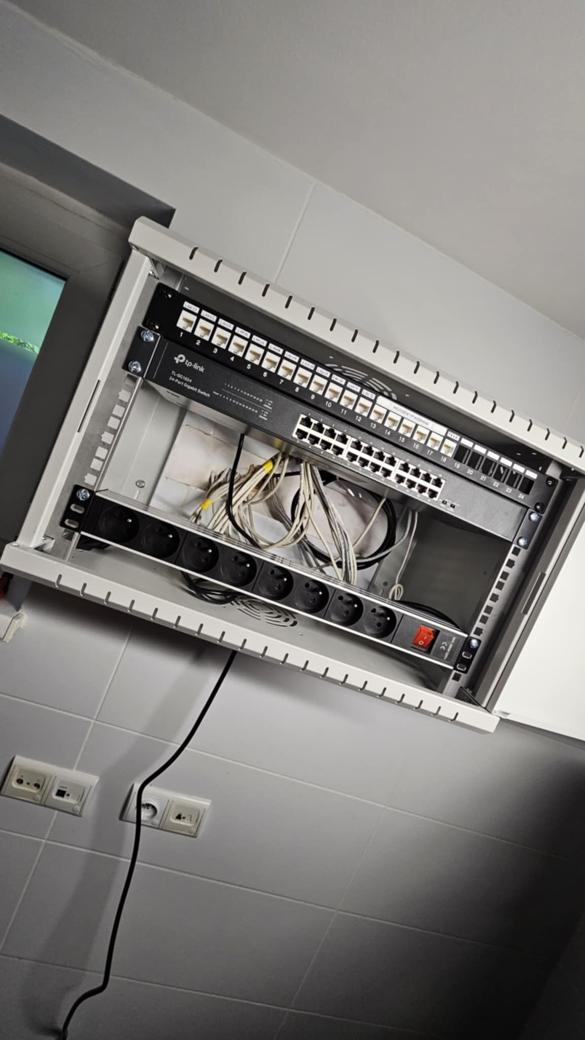 Szafa teleinformatyczna z patch panelem TP-Link, switch Gigabit i listwą zasilającą, zamontowana na ścianie z szarymi płytkami. Widoczne kable.