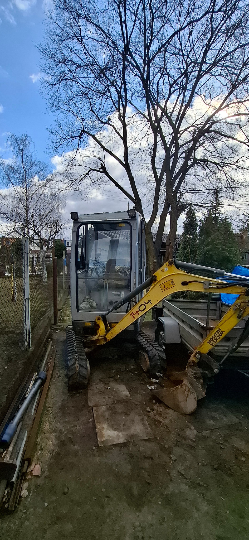 Żółto-szara mini koparka Wacker Neuson 1404 na gąsienicach, zaparkowana obok przyczepy na działce, z drzewami w tle i błękitnym niebem.