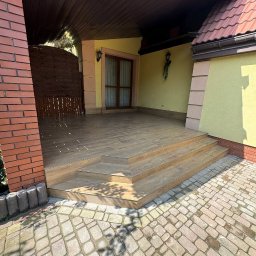 Luxe House Stepan Mukomela - Zadaszony taras z drewnianą podłogą i schodami, wykończony cegłą i kamienną kostką brukową. Widoczne detale wykończenia i oświetlenie zewnętrzne.