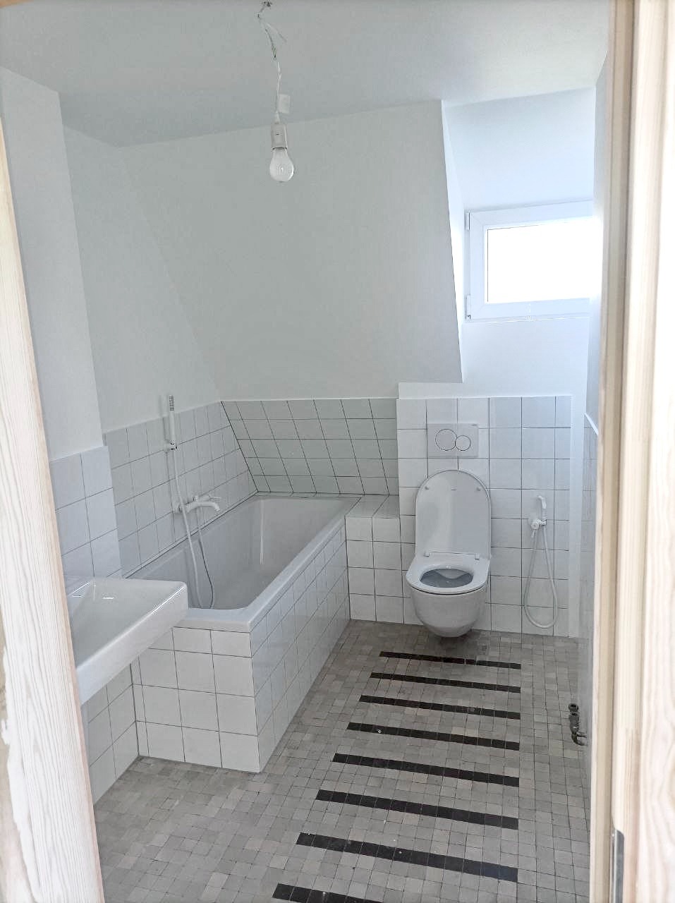 Biała łazienka na poddaszu z wanną, umywalką i toaletą. Uwagę przykuwają mozaikowe płytki podłogowe z ciemnymi pasami. Widok przez otwarte drzwi.