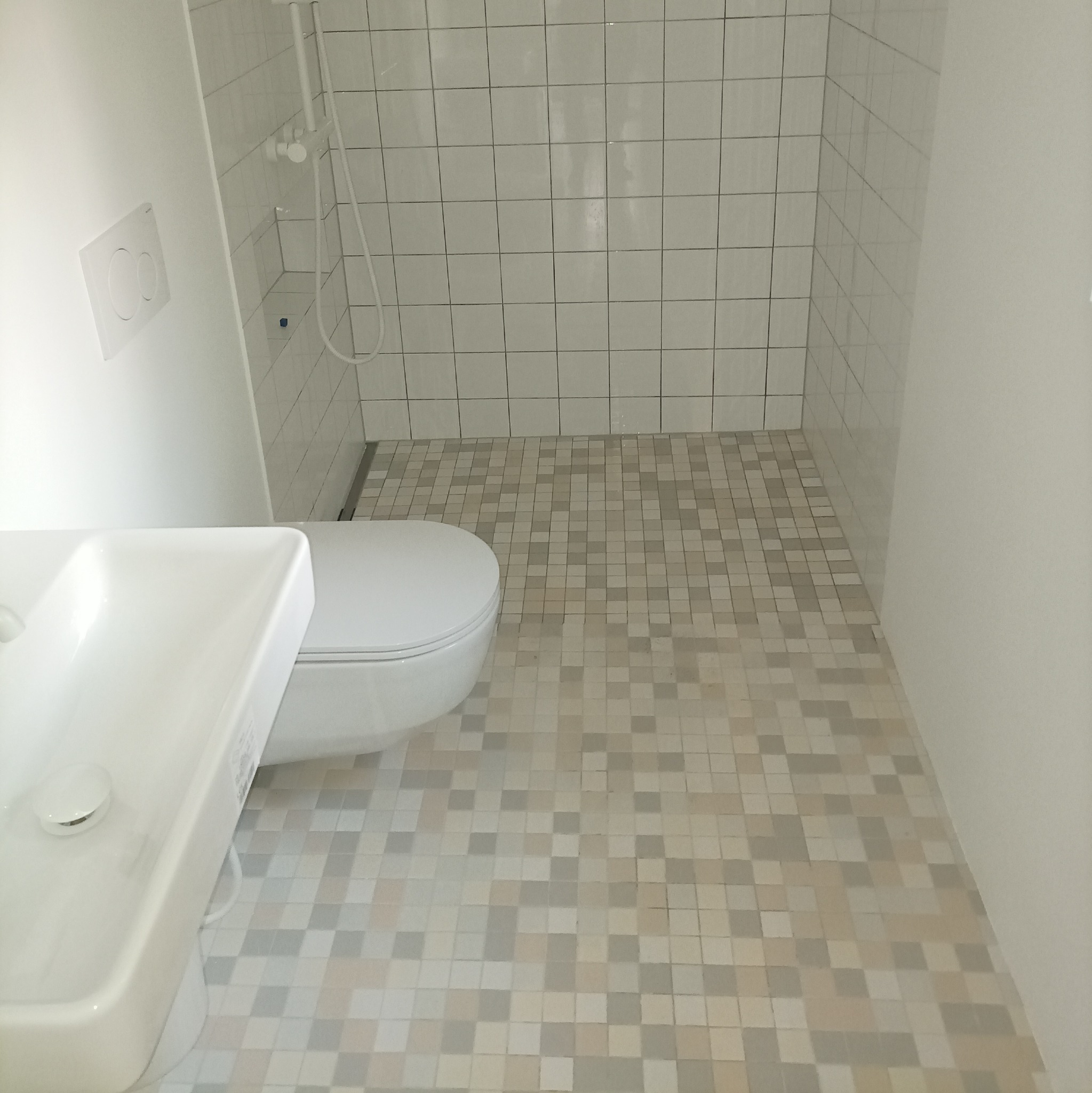 Minimalistyczna łazienka z mozaikową podłogą w odcieniach szarości i beżu, białą ceramiką sanitarną i kabiną prysznicową wyłożoną białymi, kwadratowymi płytkami.