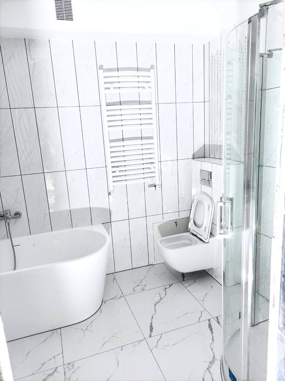 Biała łazienka z kabiną prysznicową, wanną i grzejnikiem drabinkowym. Płytki imitujące marmur z czarnymi fugami, podwieszana toaleta z otwartą klapą.