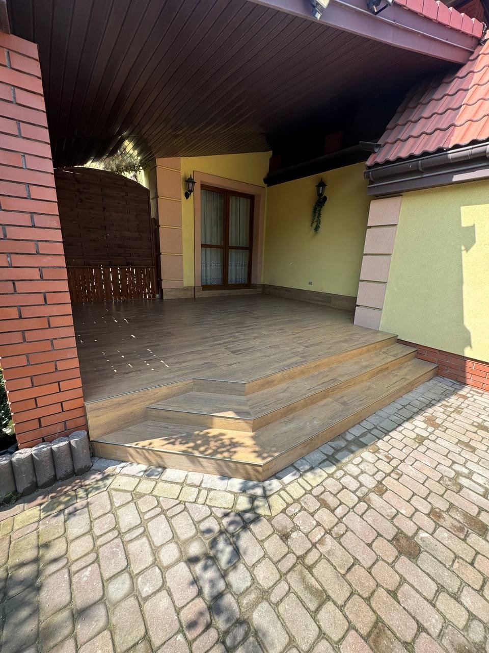 Zadaszony taras z drewnianą podłogą i schodami, wykończony cegłą i kamienną kostką brukową. Widoczne detale wykończenia i oświetlenie zewnętrzne.