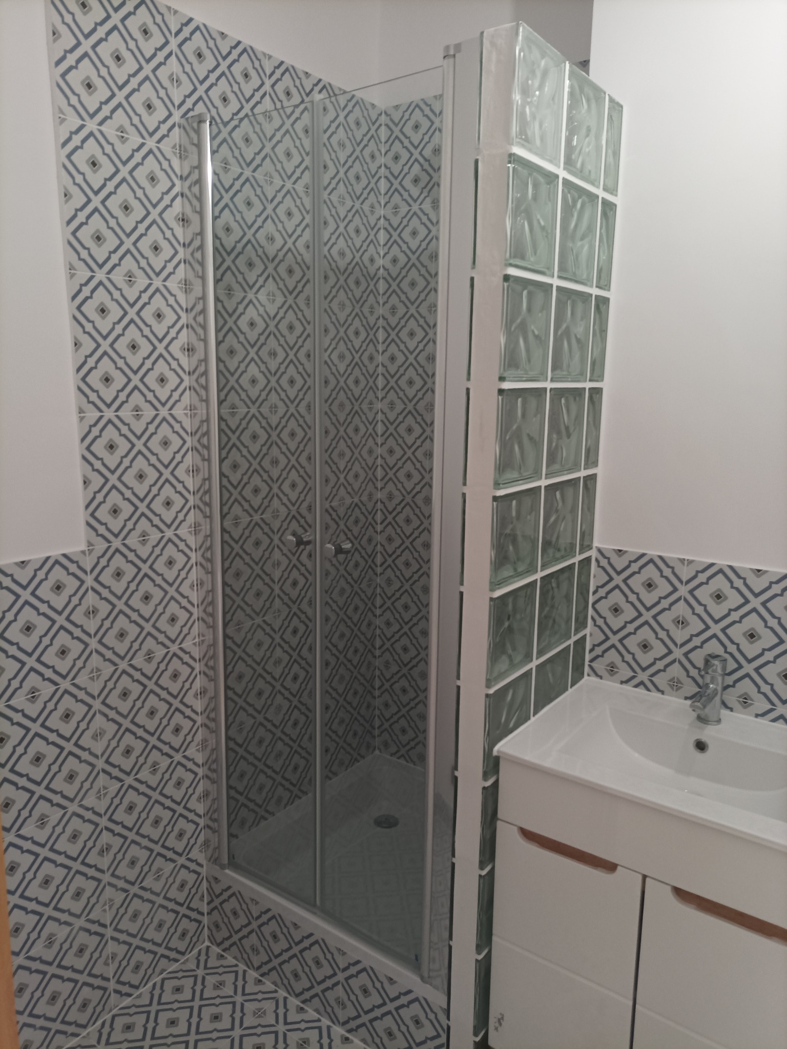 Wykończona łazienka z kabiną prysznicową, umywalką z szafką i ścianą z luksferów. Ściany wyłożone płytkami w biało-niebieskie wzory geometryczne.