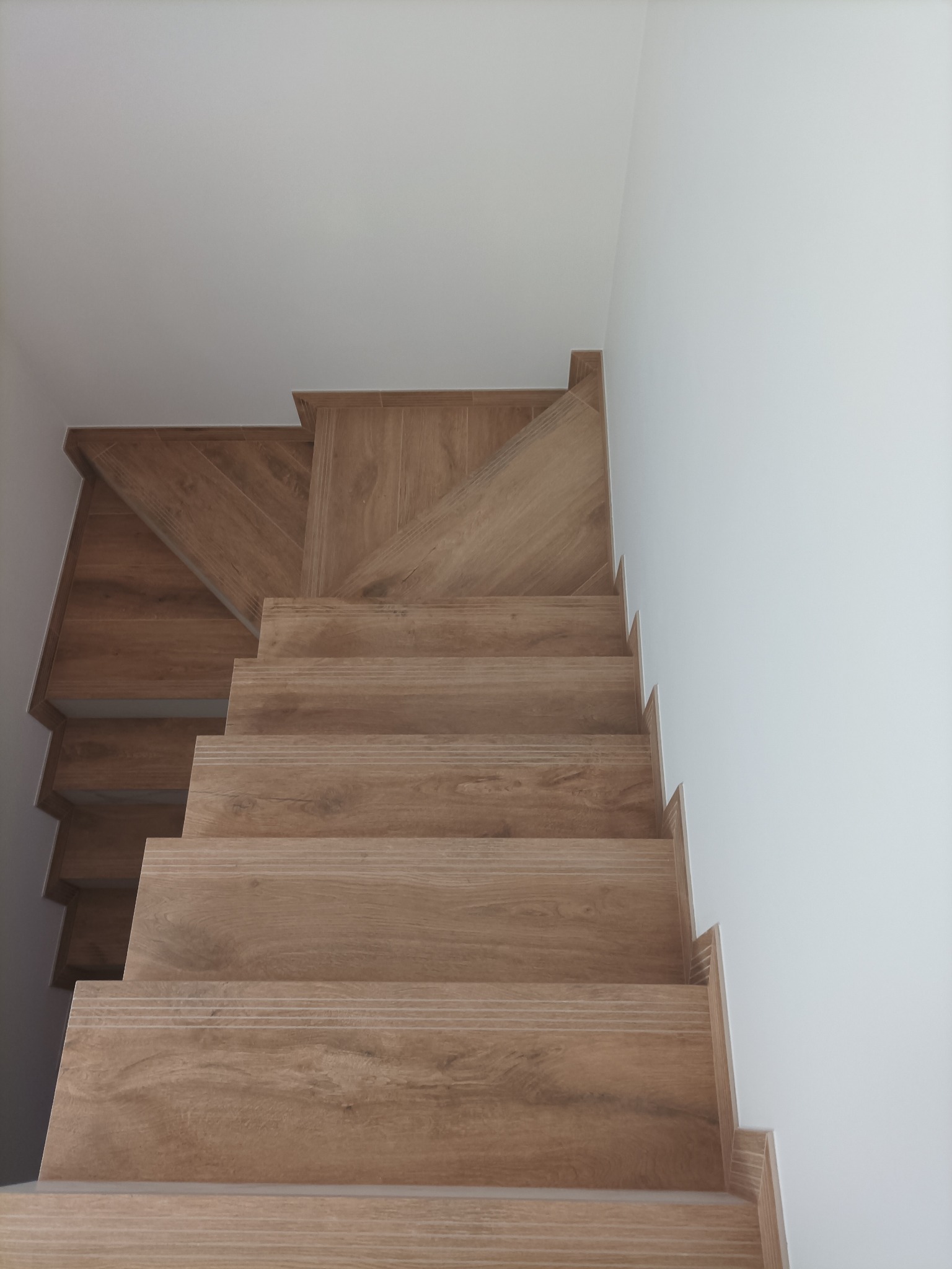 Drewnopodobne płytki na schodach wewnętrznych, widok z dołu. Stopnie wykończone z dbałością o detale, jasne ściany. Minimalistyczny styl wykończenia wnętrza.