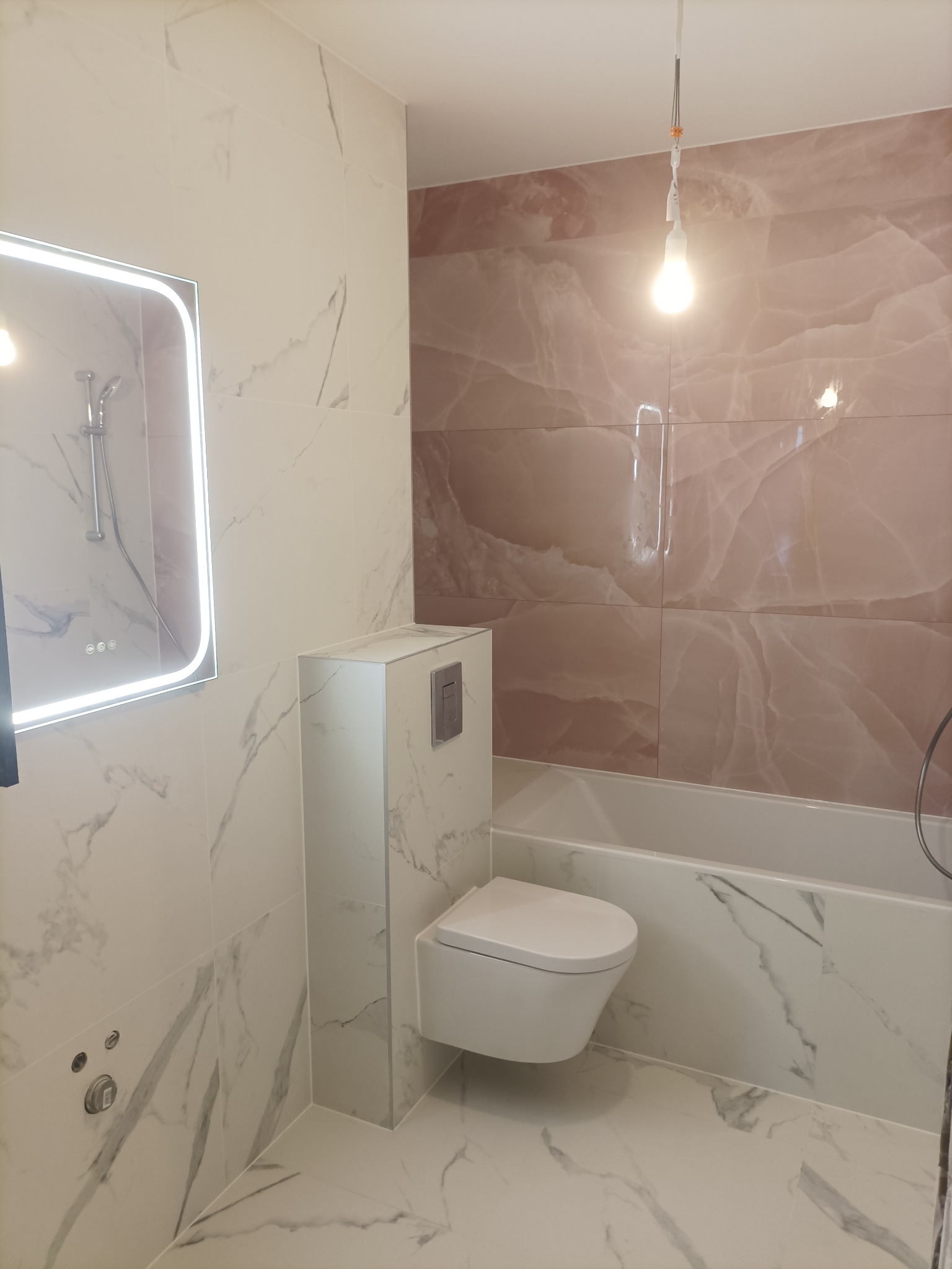 Wykończona łazienka z płytkami imitującymi marmur i onyks, podwieszana toaleta, lustro z podświetleniem LED i widoczna wanna. Całość w jasnej tonacji.