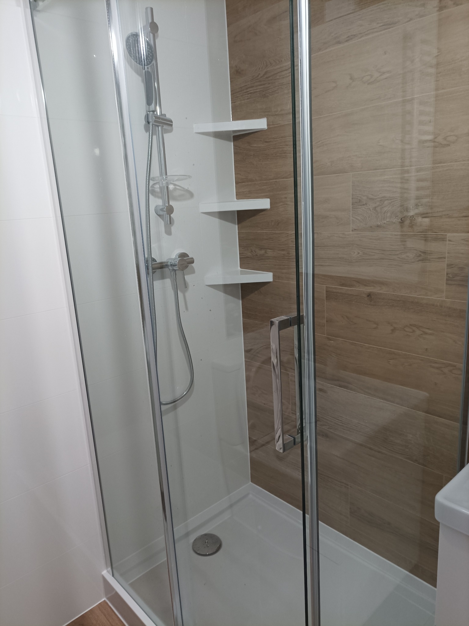 Nowoczesna kabina prysznicowa z chromowaną armaturą i półkami, wykończona białymi i drewnopodobnymi płytkami, widoczna z perspektywy wnętrza łazienki.