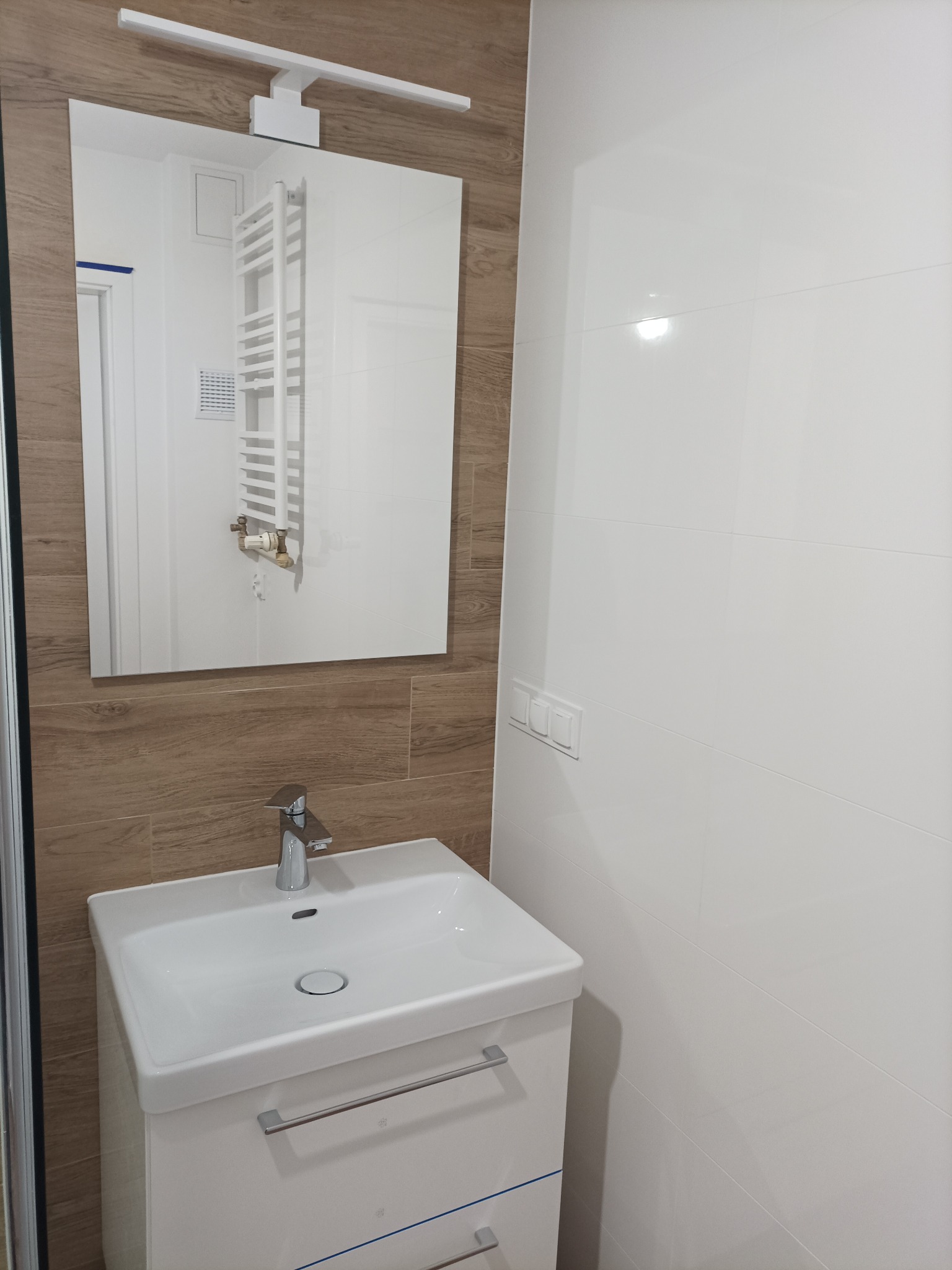 Nowoczesna łazienka po remoncie: biała umywalka z szafką, lustro z oświetleniem, białe i drewnopodobne płytki na ścianach, widoczne detale wykończenia.