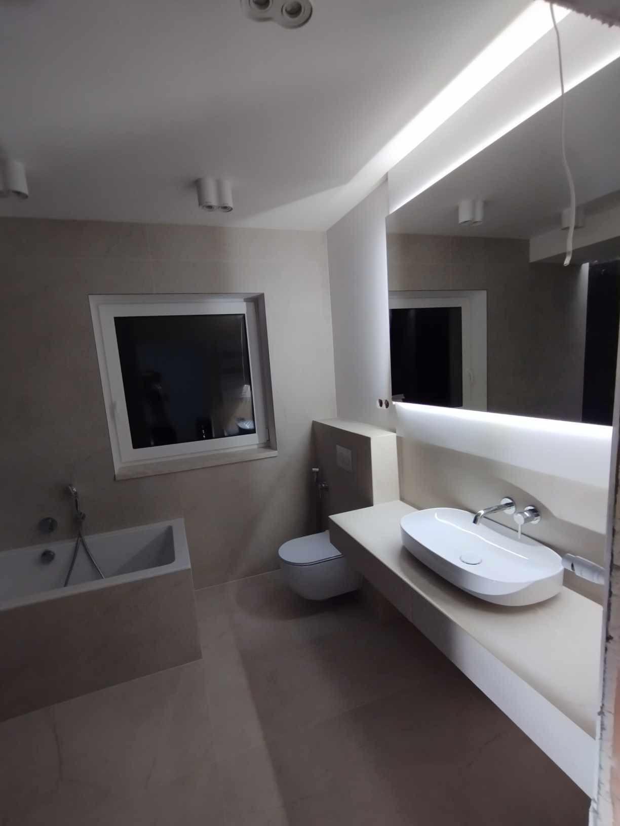 Nowoczesna łazienka w beżowej tonacji z podświetlanym lustrem, owalną umywalką i minimalistyczną armaturą. Widoczna wanna i toaleta w zabudowie.