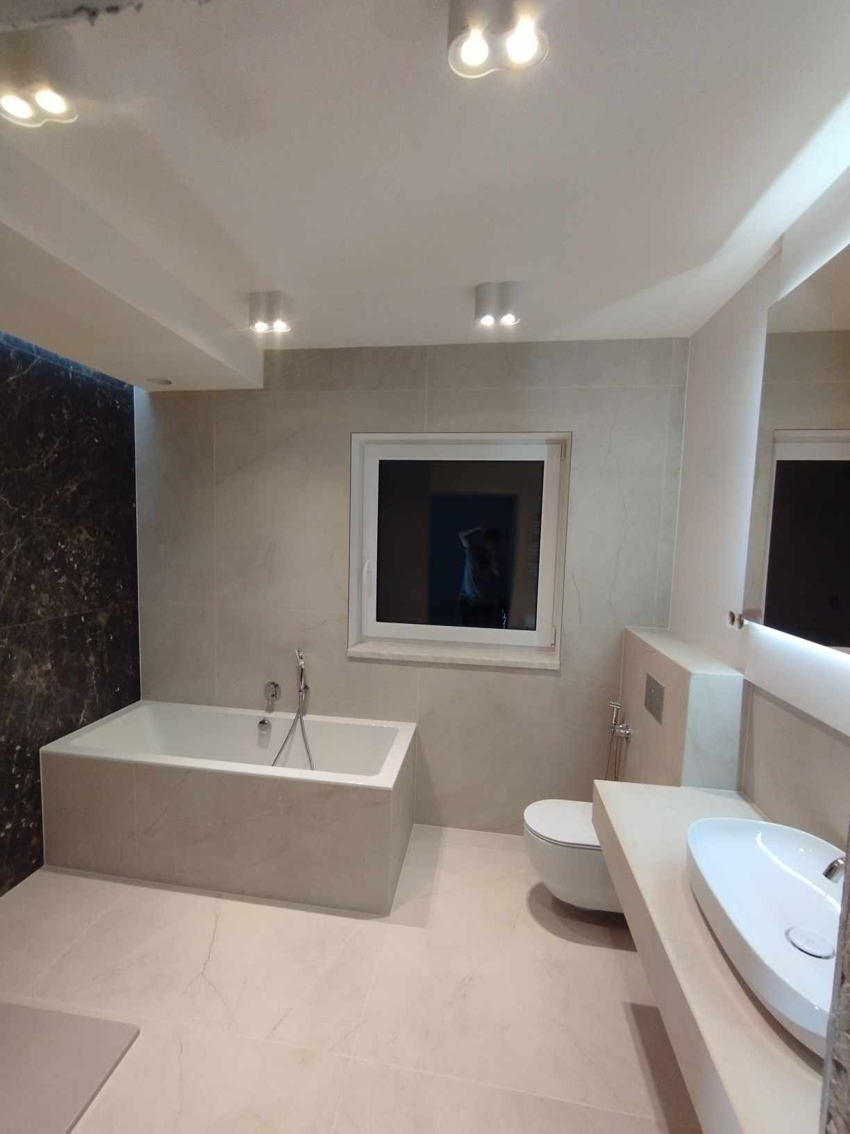 Nowoczesna łazienka w beżach: wanna obudowana płytkami, toaleta podwieszana, umywalka nablatowa i czarna ściana z marmuru. Oświetlenie punktowe.