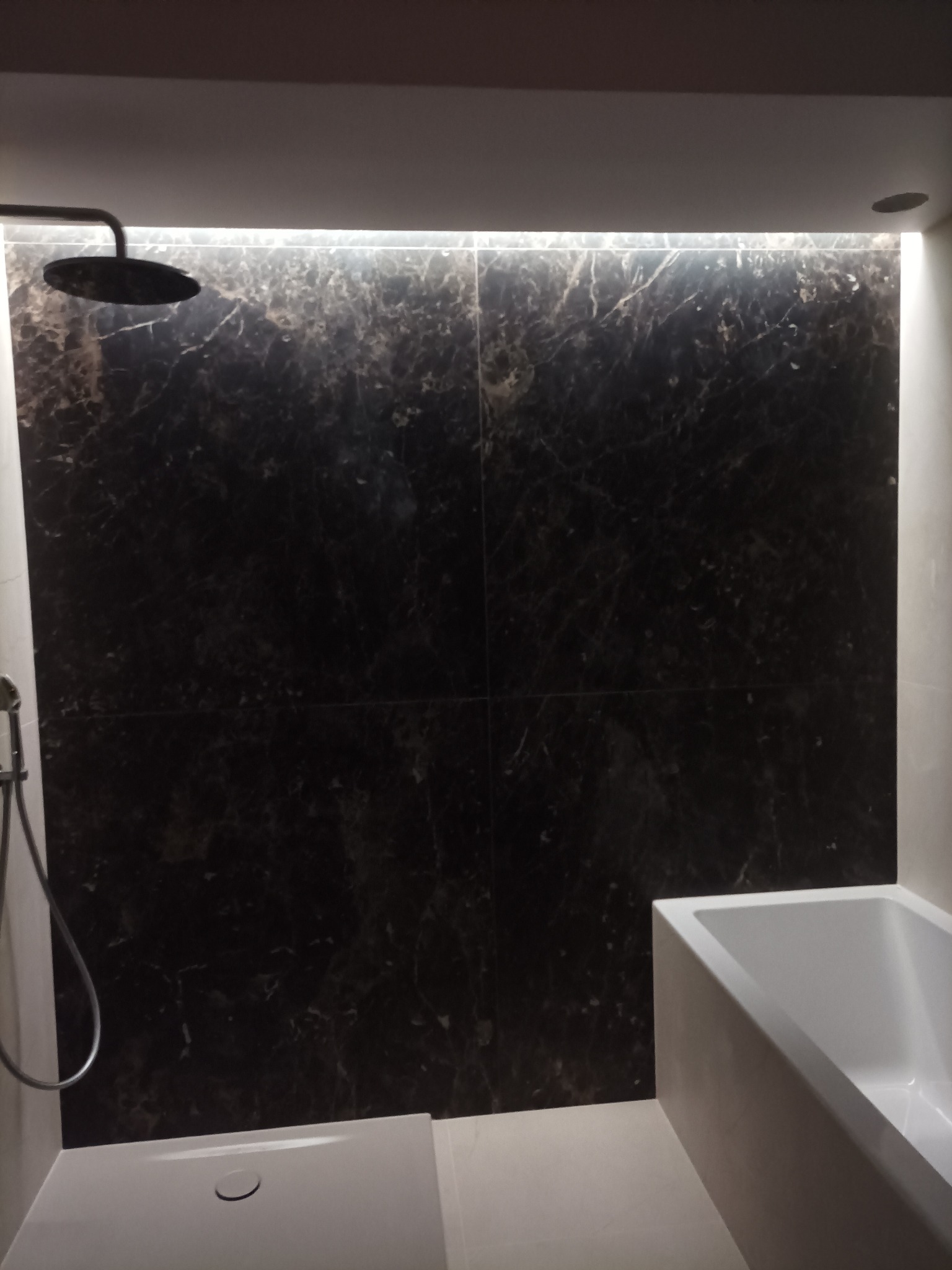 Nowoczesna łazienka z czarną, marmurkową ścianą podświetloną LED, deszczownicą i białą wanną. Minimalistyczny design, eleganckie wykończenie.