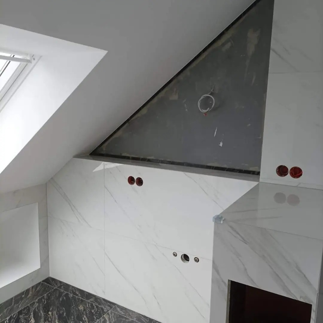 Łazienka na poddaszu ze skośnym oknem dachowym i marmurowymi płytkami. Widoczne otwory pod instalacje elektryczne i fragment surowego sufitu.