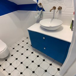 Slavik Kyselov - Wykończona łazienka z biało-czarną podłogą, niebieską szafką pod umywalką i lustrem zabezpieczonym folią. Widoczne złote elementy armatury i białe heksagonalne płytki.