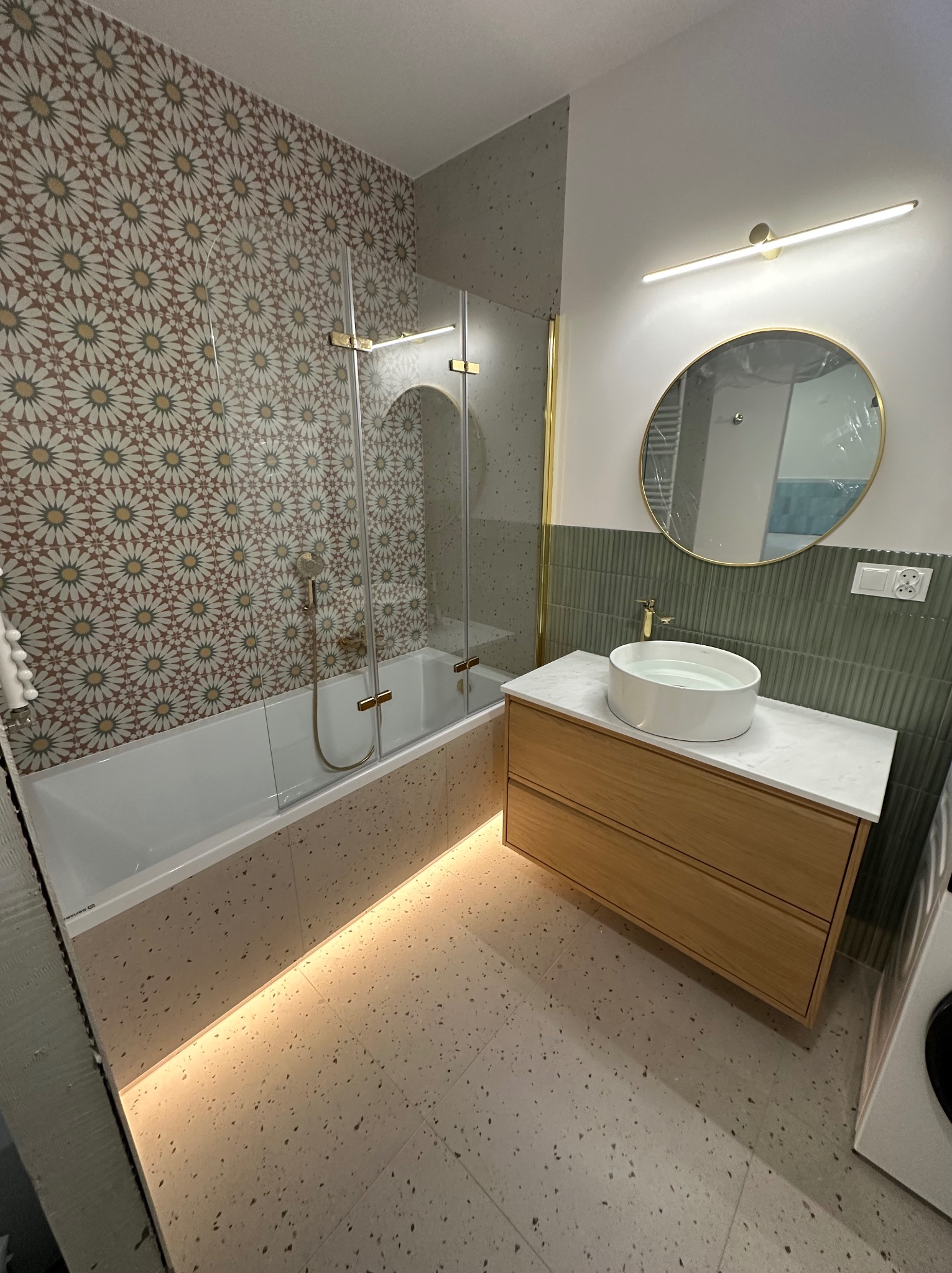 Nowoczesna łazienka z wanną za szklanym parawanem, umywalką na drewnianej szafce i dekoracyjnymi płytkami w odcieniach beżu i brązu. Oświetlenie LED podkreśla detale.