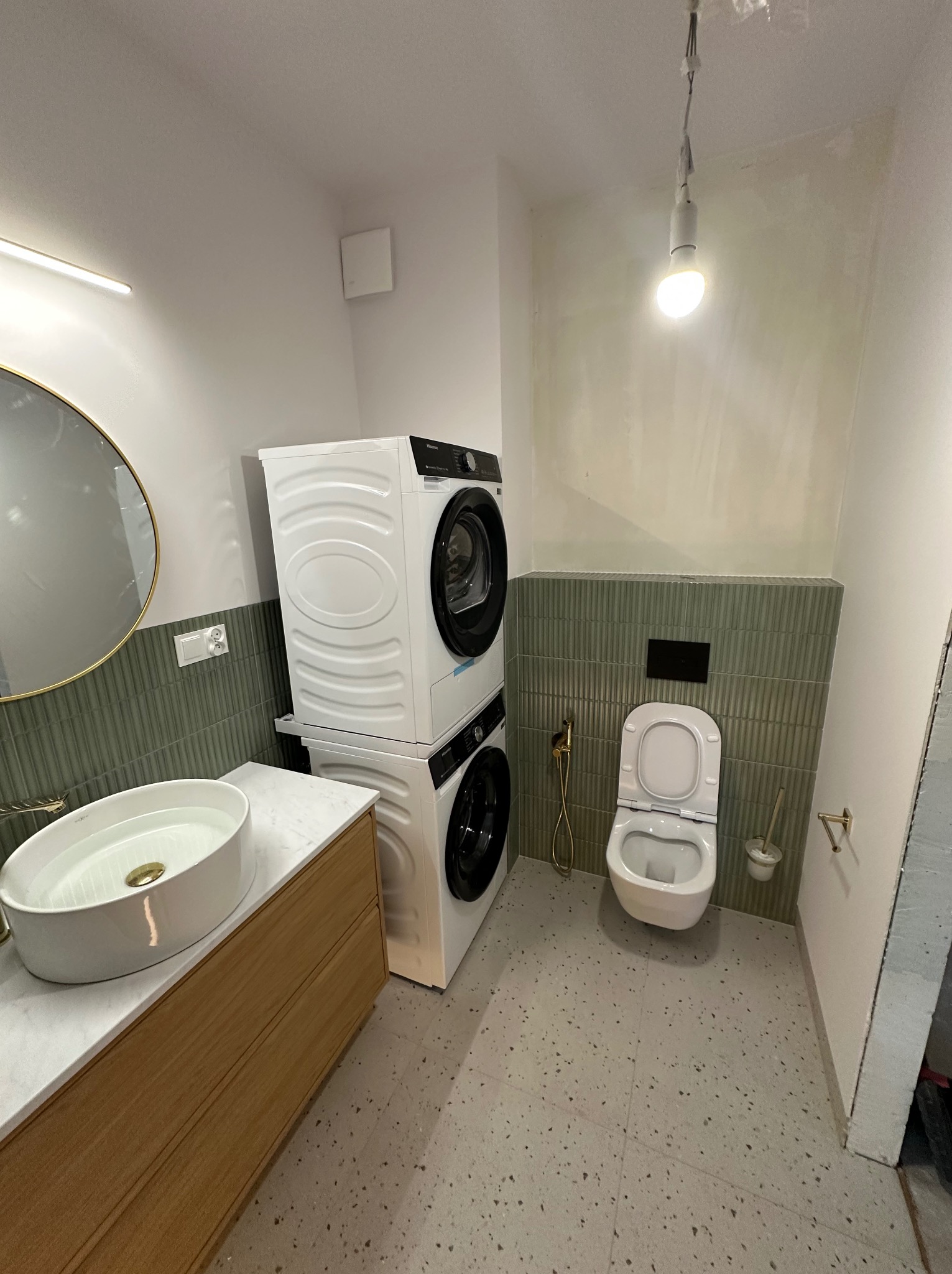 Nowoczesna łazienka z pralko-suszarką w pionie, umywalką na blacie, toaletą wiszącą i zielonymi płytkami. Minimalistyczny design, szare płytki podłogowe.